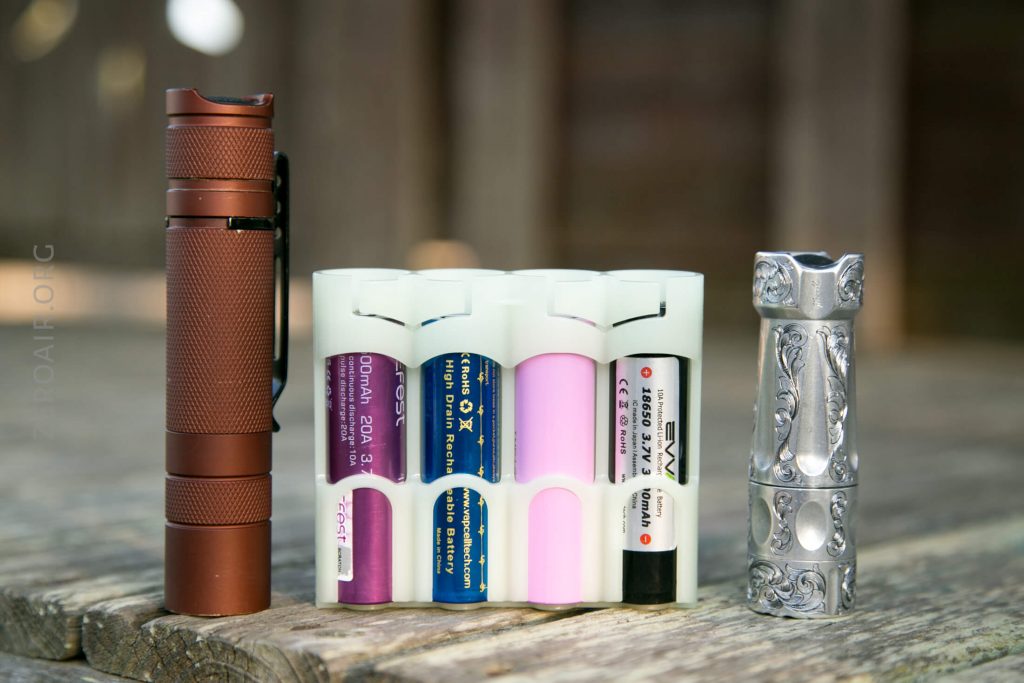 A brown flashlight, a white battery holder containing four cylindrical batteries of various colors, and a silver, ornately designed flashlight are arranged side by side on a wood surface.