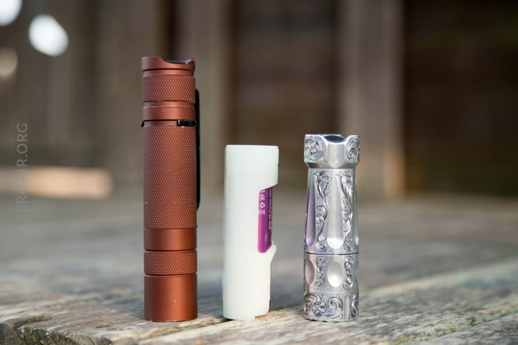 Three cylindrical objects stand upright on a wooden surface: a brown textured flashlight, a white translucent battery case with a visible battery inside, and a small engraved silver flashlight. The background is blurred.