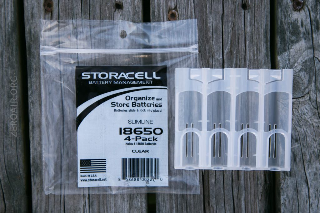 A clear plastic package labeled “Storacell 18650 4-Pack” sits on a wooden surface next to four transparent plastic battery holders designed to fit 18650 batteries.