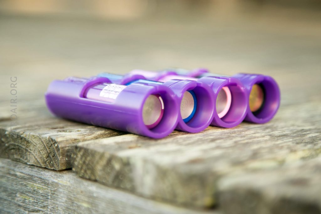 Four purple inhalers are lined up side by side on a wooden surface, viewed from the side with their mouthpieces facing forward. The background is softly blurred.