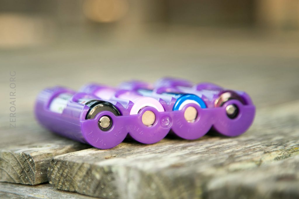 Four AA batteries with purple plastic battery holders are placed side by side on a wooden surface, photographed close up with a shallow depth of field.