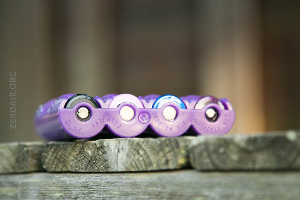 Four purple cylindrical batteries are placed horizontally side by side on a weathered wooden surface. The sides show branding and markings, including D.M. PATENTED and MADE IN USA. Background is blurred.