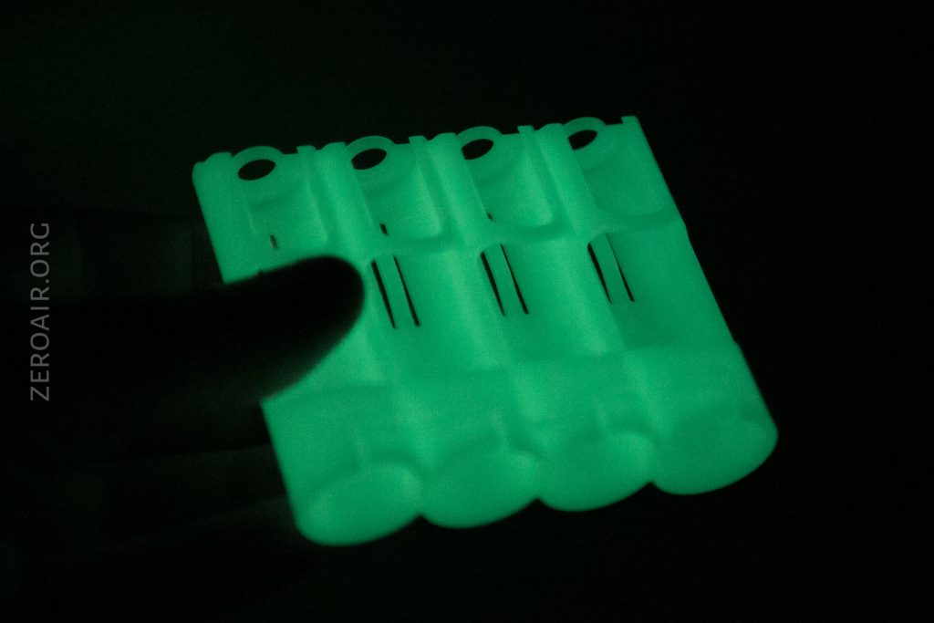 A hand holds a semi-transparent, rectangular object that glows green in the dark. The object has four cylindrical slots, likely for batteries. The text ZEROAIR.ORG is visible on the left side.