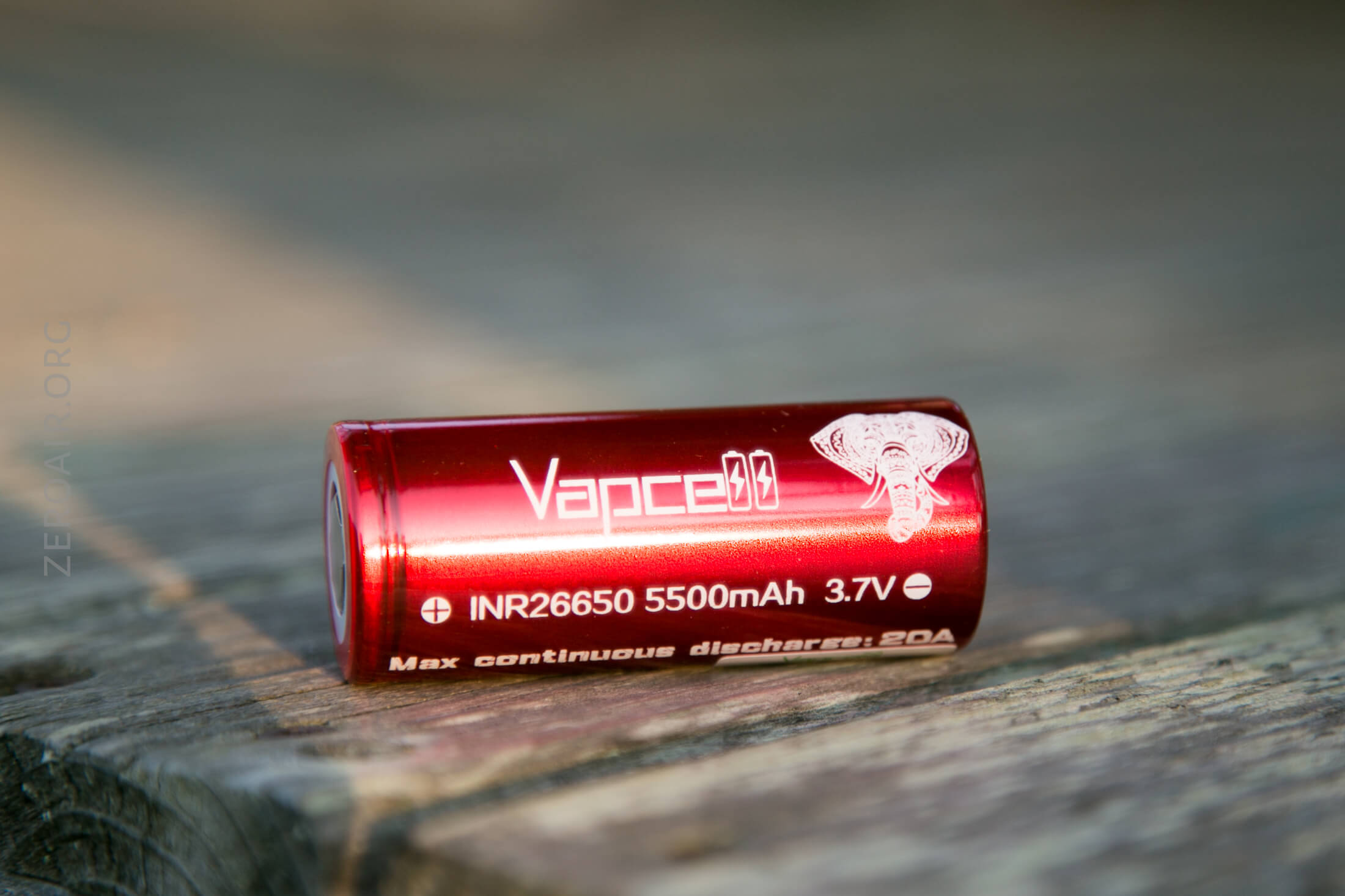 A red cylindrical battery labeled “Vapcell” with specifications INR26650, 5500mAh, 3.7V, max continuous discharge 20A, and an elephant logo, resting on a wooden surface.