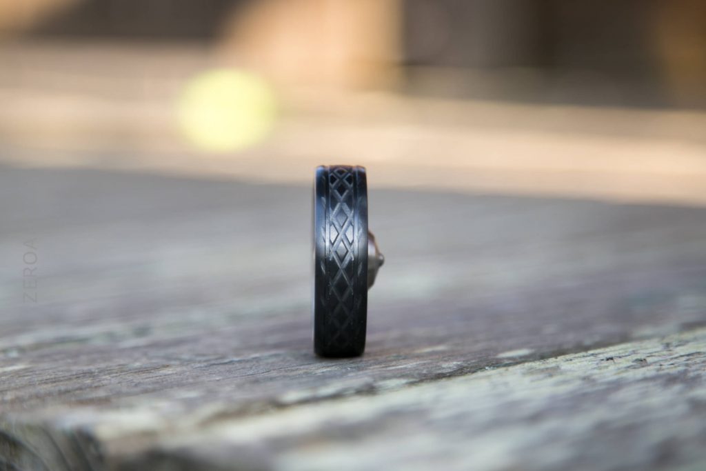 A close-up of a small black wheel with a textured pattern is standing upright on a weathered wooden surface, with the background softly blurred.