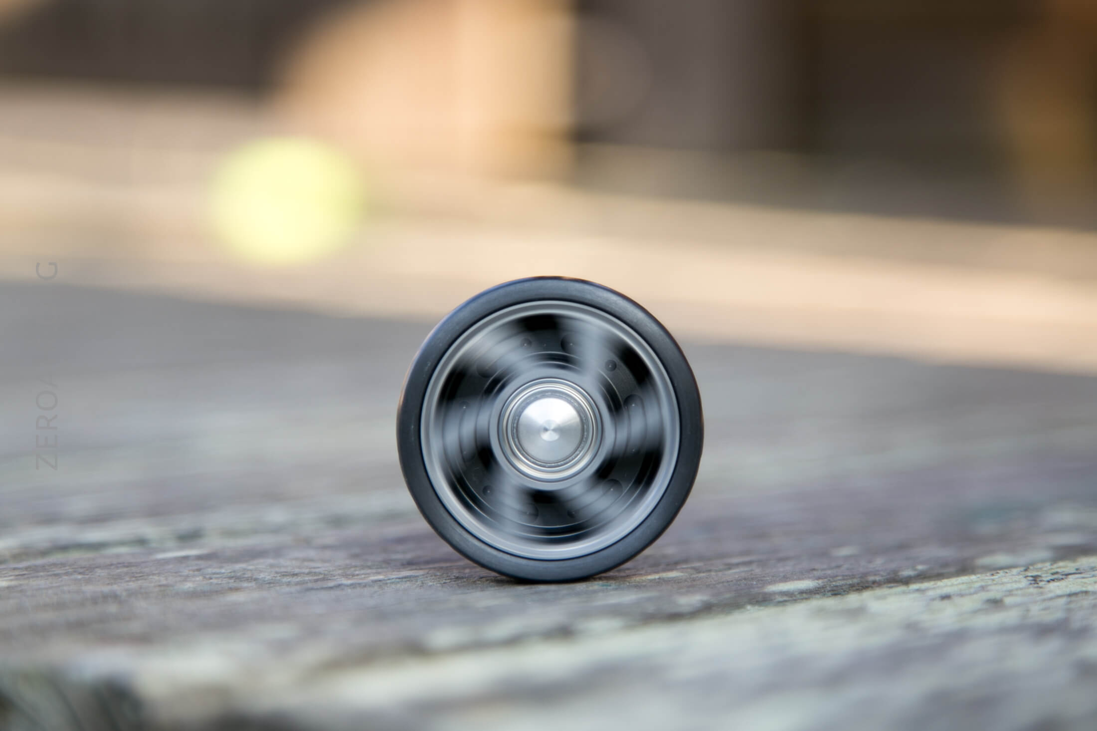 A close-up of a spinning yo-yo, photographed head-on while resting on a wooden surface. The yo-yo’s motion creates a blurred, circular pattern in the center. The background appears out of focus.