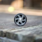 A black and silver yo-yo is standing upright on a weathered wooden surface outdoors, with a blurry yellow-green object in the background.