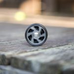 A single black yo-yo with a silver center rests upright on weathered wooden steps, with a blurred background and a faint yellow object in the distance.
