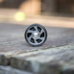 A black yo-yo with a metallic center is resting upright on a weathered wooden surface, with blurred background elements out of focus.