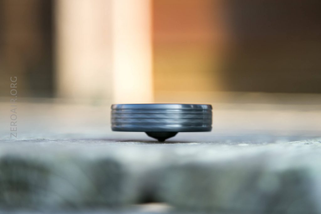 A close-up view of a metallic spinning top balanced on a rough surface, with a blurred background featuring warm tones. The words ZEROAIR.ORG are faintly visible on the left side.