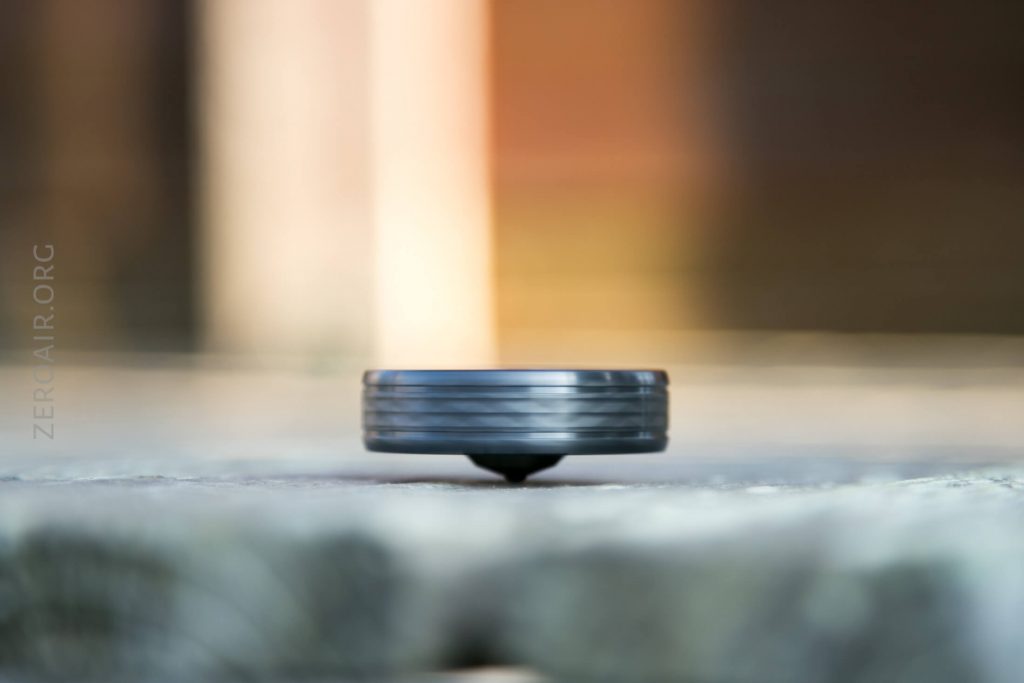 A small, dark spinning top balances on its pointed tip on a flat surface, with a blurred background of soft brown and green tones. The word ZERGAIR.ORG is faintly visible along the left side.