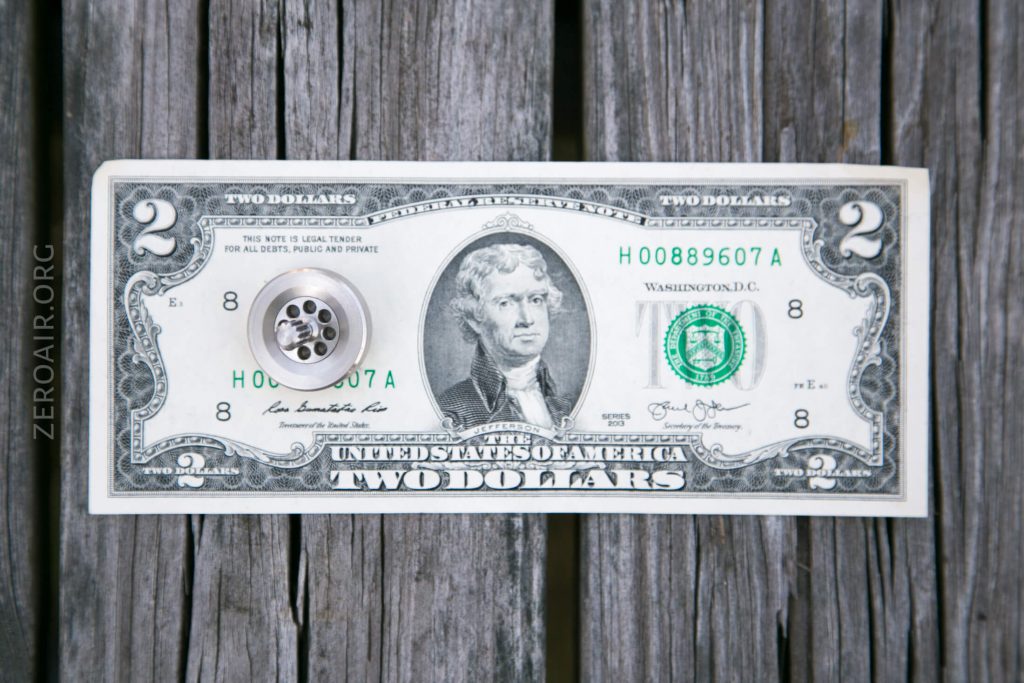 A U.S. two-dollar bill lies flat on a wooden surface with a silver metal button or knob placed over Thomas Jeffersons portrait in the center.