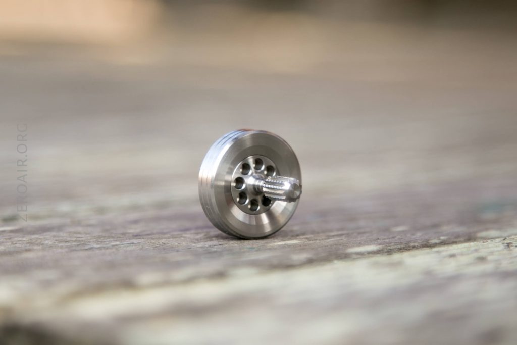 A small, metallic gear with holes and a central shaft is standing upright on a blurred, wooden surface.