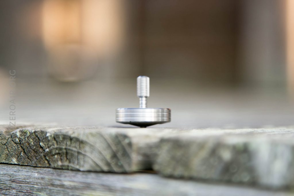A silver spinning top balances upright on a weathered wooden surface, with a blurred background in neutral tones.