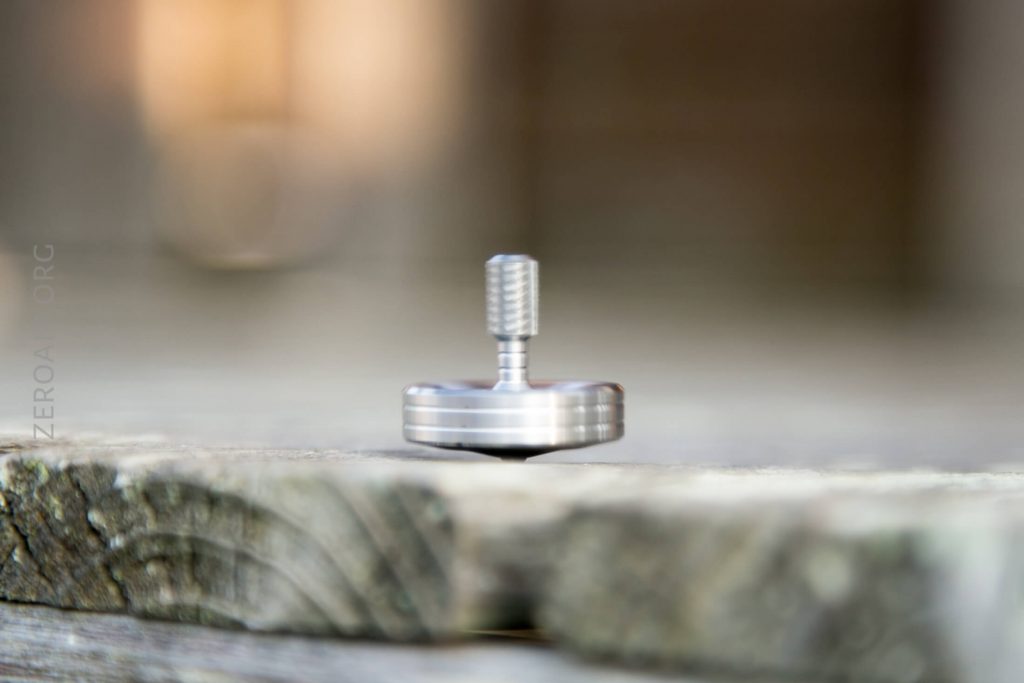 A metal spinning top is balanced and spinning on a weathered wooden surface, with the background blurred and out of focus.