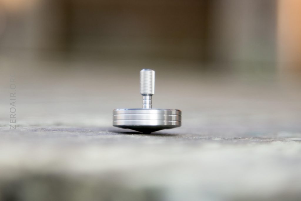 A close-up side view of a silver metal spinning top balanced on its tip on a light, textured surface, with a blurred background.