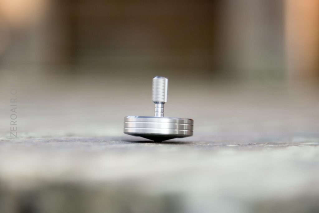 A close-up photo of a silver metal spinning top balanced on its tip on a flat surface, with the background blurred.