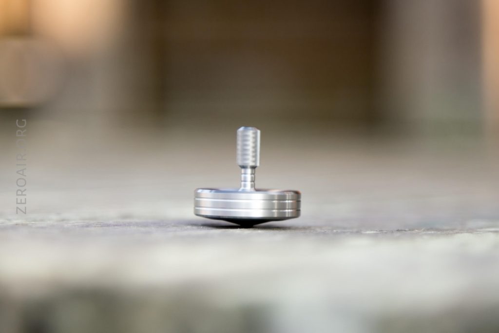A close-up of a metallic spinning top standing upright on a flat surface, with a blurred background and soft lighting.