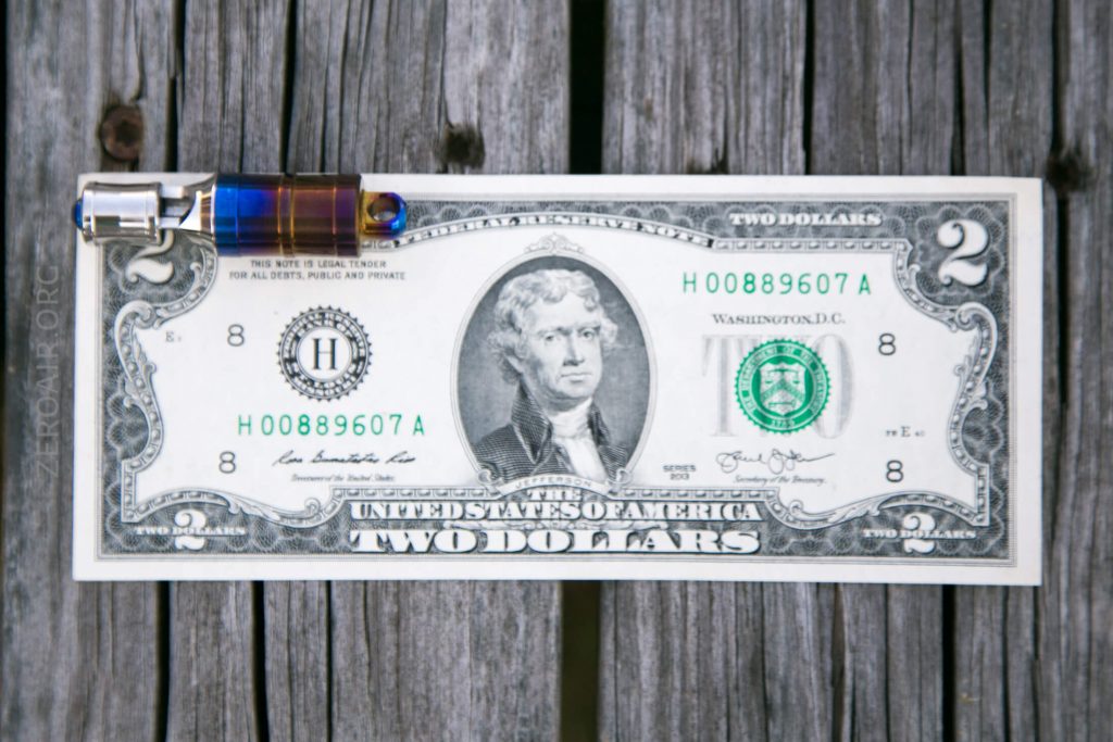 A two dollar bill lies flat on a weathered wooden surface with a small pen placed horizontally across the top left side of the bill.