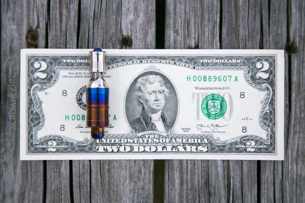 A vape cartridge filled with liquid is placed vertically on top of a two-dollar bill, which rests on a weathered wooden surface.