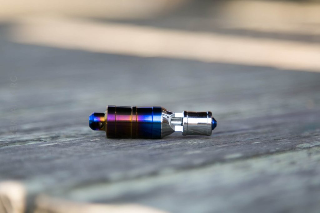 A small, metallic, cylindrical object with a gradient color from purple to blue and silver, rests on a weathered wooden surface outdoors.