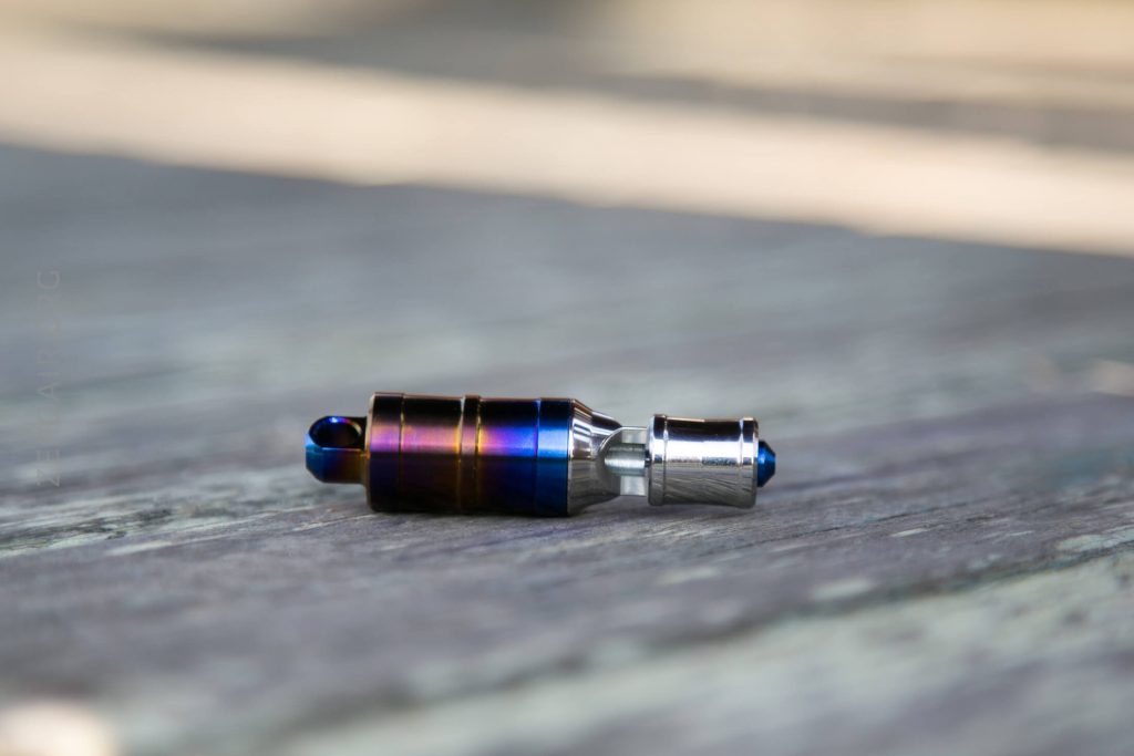 A small metallic object with a cylindrical shape and a gradient blue and silver finish lying on a wooden surface. The object has a rounded end and a narrow, silver tip.