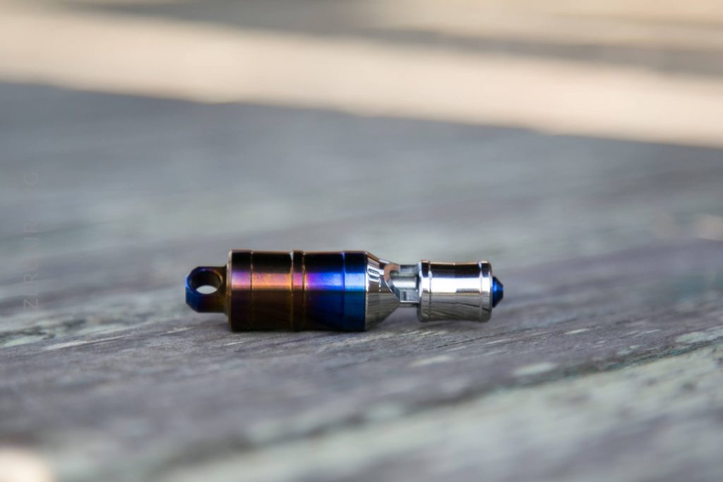 A small metallic object with a cylindrical shape and a gradient blue and brown finish lies on a weathered wooden surface. The item appears to have a keyring hole on one end and a metallic connector on the other.