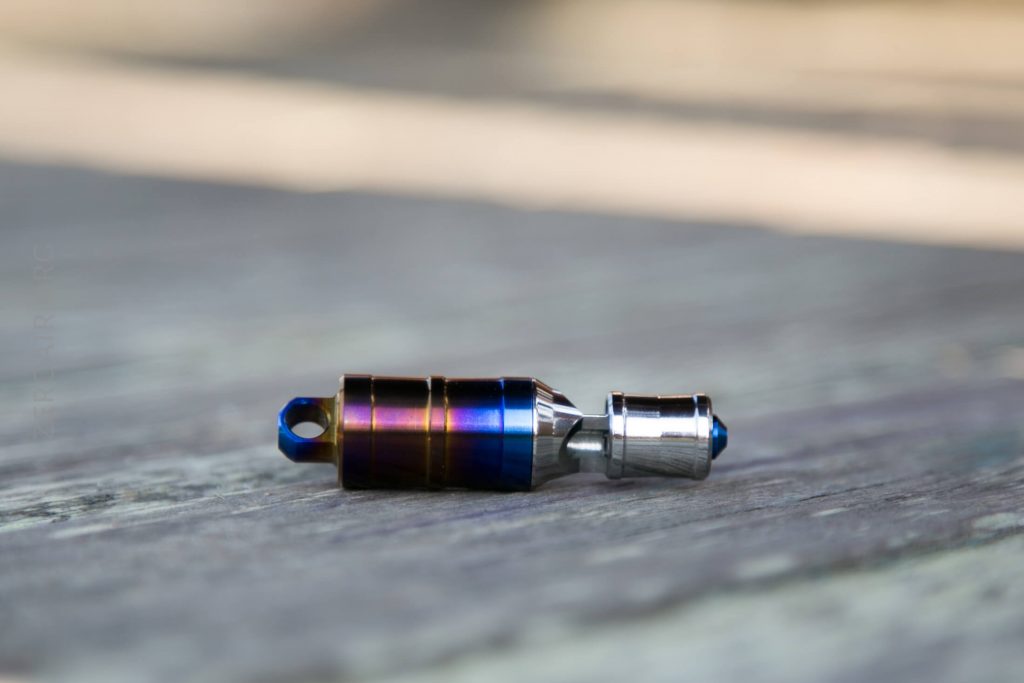 A small, metallic, cylindrical object with blue, purple, and silver sections rests on a wooden surface. The background is out of focus.