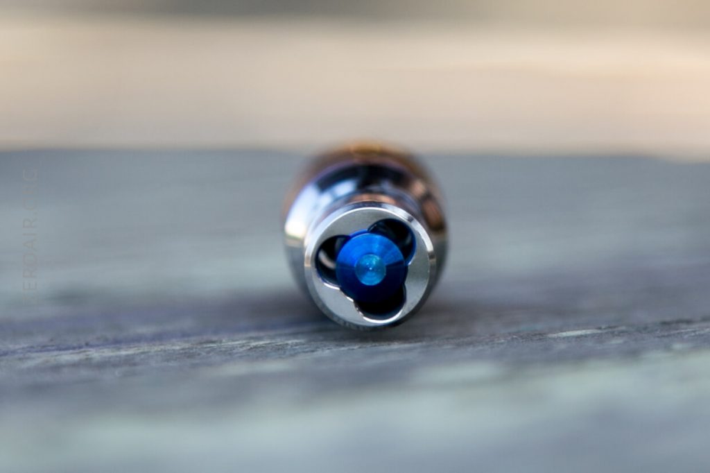 Close-up of a metallic cylindrical object with a blue tip, viewed from the front and resting on a wooden surface. Background is blurred.