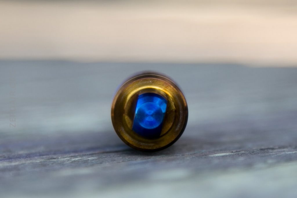 Close-up of the base of a bullet lying on its side, with a metallic casing and a central blue primer, photographed on a blurred wooden surface.