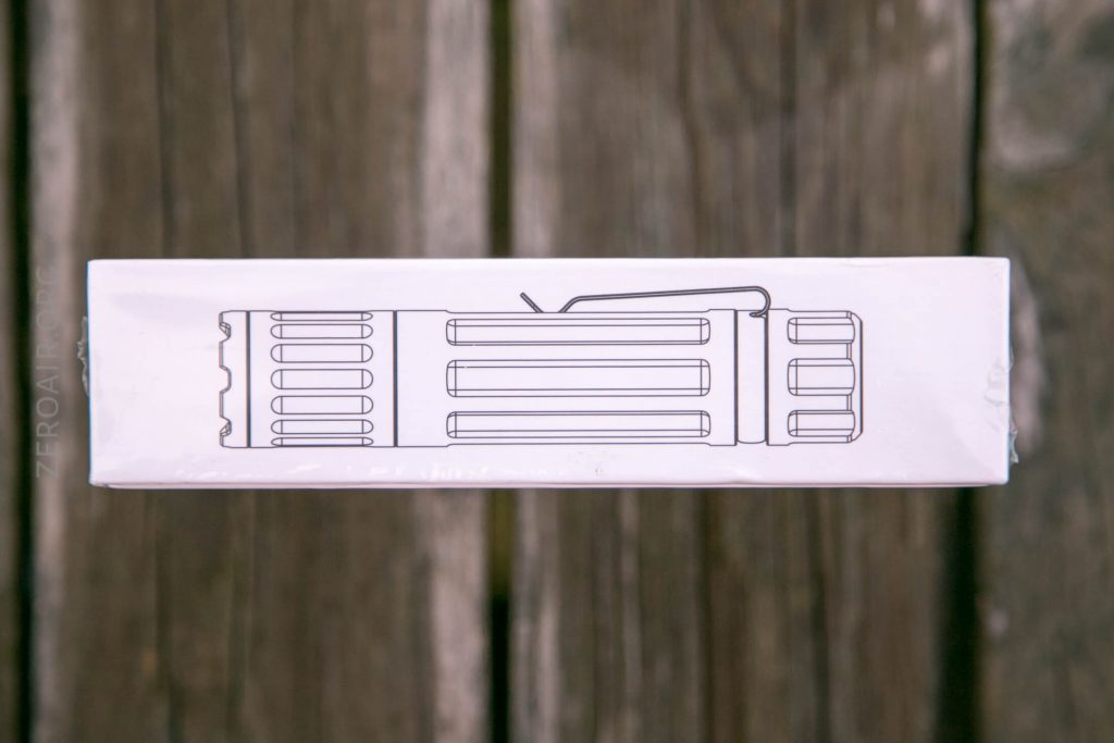 A white rectangular box with a black line drawing of a flashlight on its side is placed on a wooden surface. The drawing shows the flashlight in a side profile with visible grooves and a pocket clip.