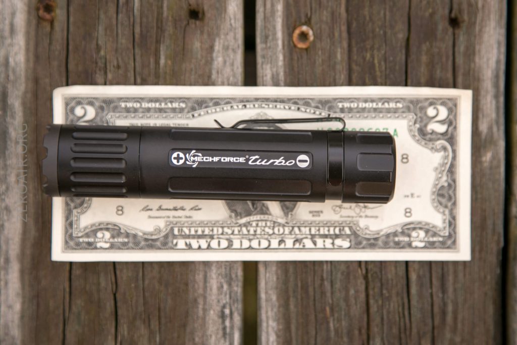 A black Mechforce Turbo flashlight is placed horizontally on top of a two-dollar bill, both resting on a weathered wooden surface.