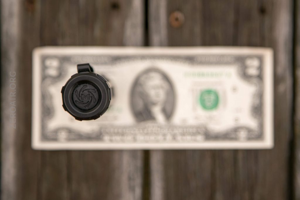 A close-up, top-down view of a black flashlight facing the camera, positioned above a two dollar bill on a wooden surface. The bill is out of focus in the background.