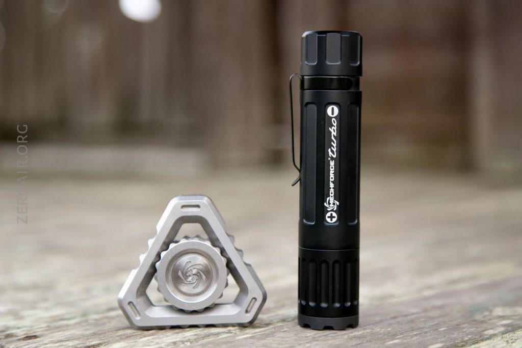 A black cylindrical flashlight stands upright next to a small, triangular metal fidget spinner with a swirl design, both placed on a weathered wooden surface with a blurred brown background.
