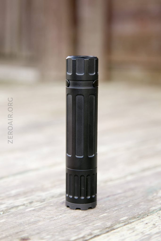 A black, cylindrical flashlight or tool stands upright on a wooden surface with a blurred brown background. The object has vertical grooves and ridges along its body. The text ZEROAIR.ORG is visible on the left side.