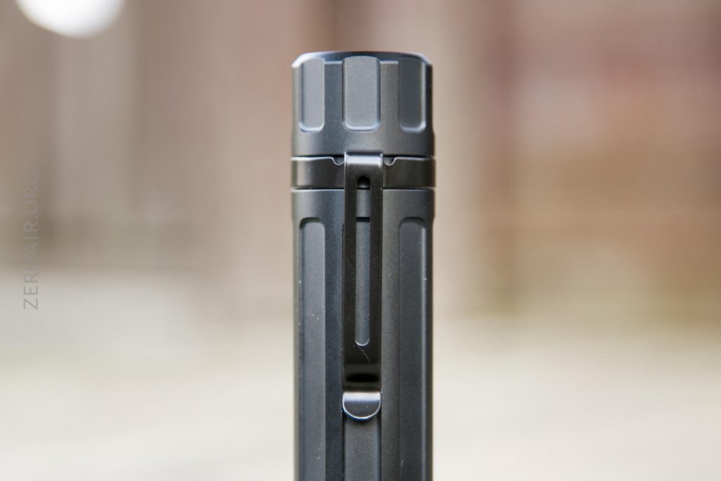 A close-up view of a black, cylindrical metal object with a textured top and a vertical clip attached to its side, set against a blurred background.