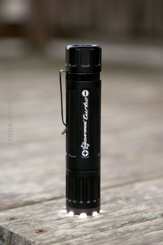 A black cylindrical flashlight with a pocket clip is standing upright on a wooden surface. The flashlight has branding and text on its side and features a ridged design on both ends.