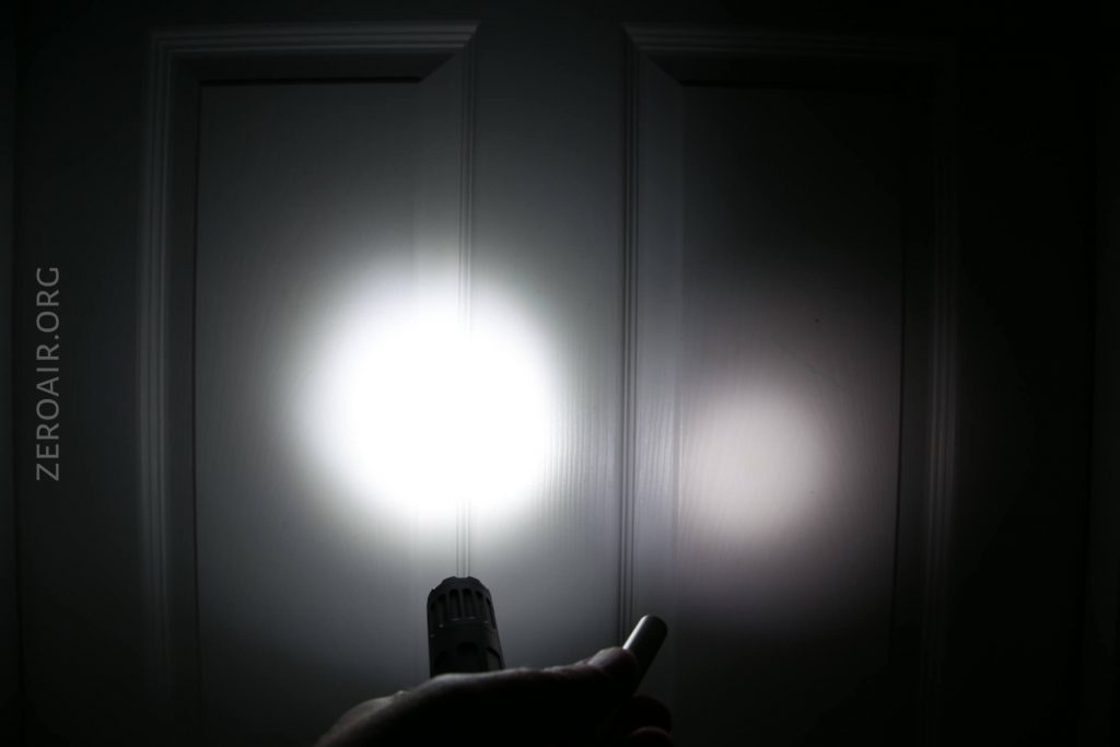 A person is shining a flashlight at a white paneled door in a dark room, creating a bright circular beam of light with a smaller, dimmer spot to the right. The word ZEROAIR.ORG appears on the left side.