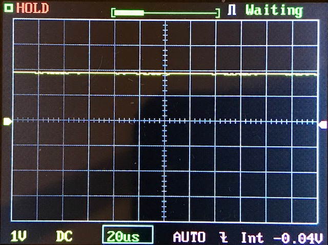 Oscilloscope screen shows a nearly flat yellow waveform along the horizontal axis, with settings at 1V/div, 20 microseconds/div, DC coupling, and a measurement reading of -0.04V. The status displays HOLD and Waiting.
