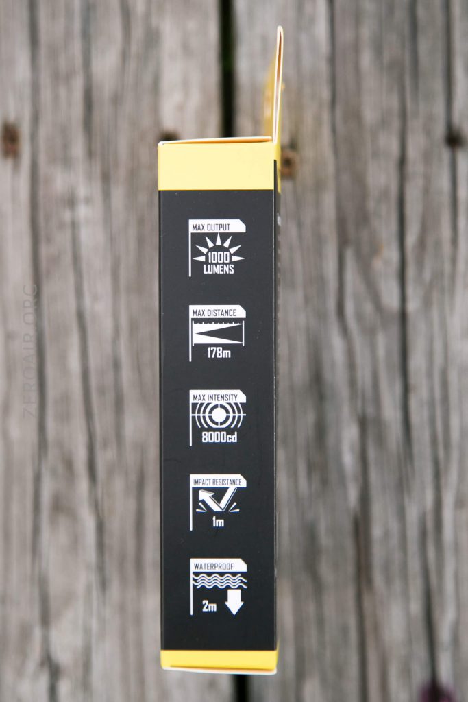 A black and yellow product box displays specifications: max output 1000 lumens, max distance 178 meters, max intensity 8000cd, impact resistance 1 meter, and waterproof to 2 meters. The background is weathered wood.