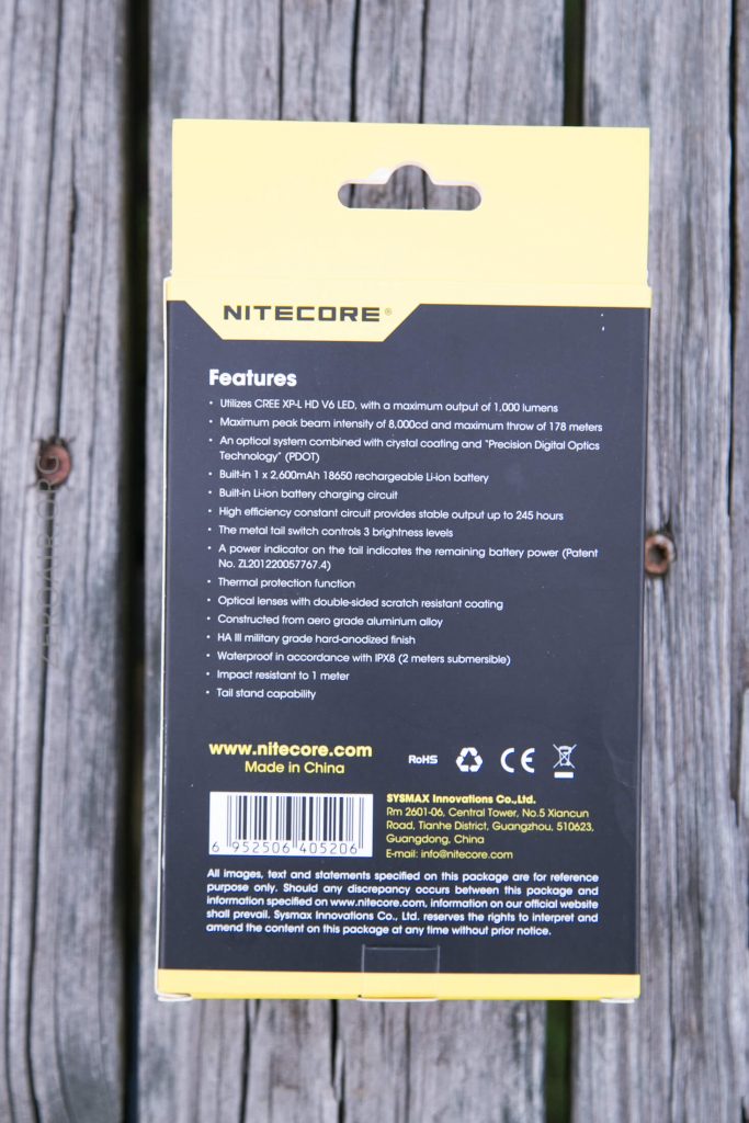 The back of a Nitecore product box showing features and specifications, including max output of 1,000 lumens, rechargeable Li-ion battery, runtime, and company information. Box background is yellow and black, set on a wooden surface.