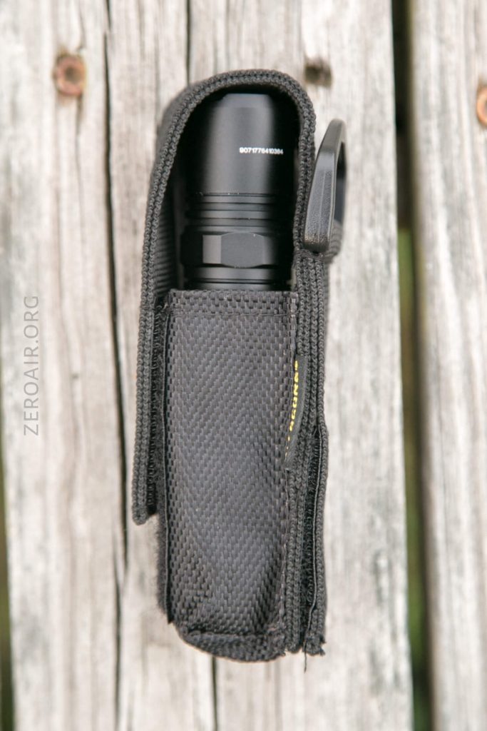 A black flashlight inside a textured black nylon holster with a hook-and-loop flap, placed on a weathered wooden surface. The text ZEROAIR.ORG is faintly visible along the left side of the image.