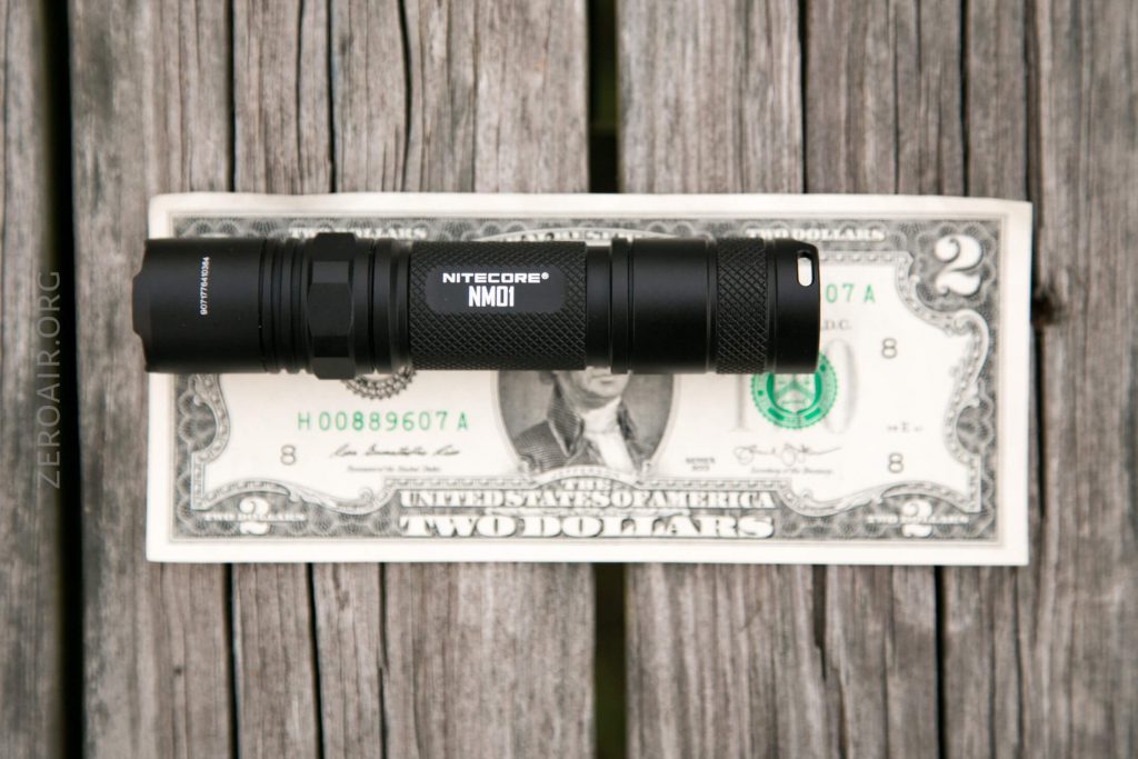 A black Nitecore NM01 flashlight is placed horizontally on top of a two-dollar bill, which is laying on a weathered wooden surface.