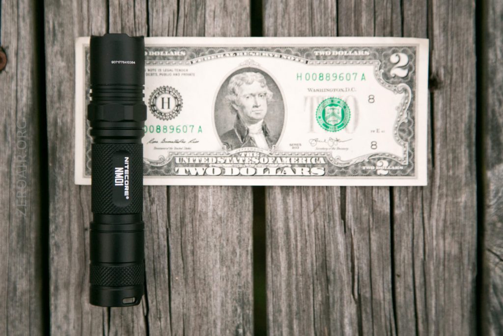 A black flashlight is placed next to a U.S. two dollar bill on a weathered wooden surface, showing their relative sizes.