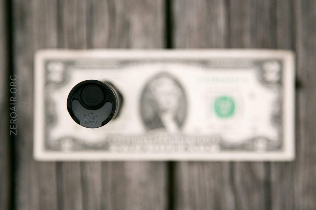 A black bottle cap with an open symbol is in focus, positioned above a blurred two-dollar bill on a wooden surface.