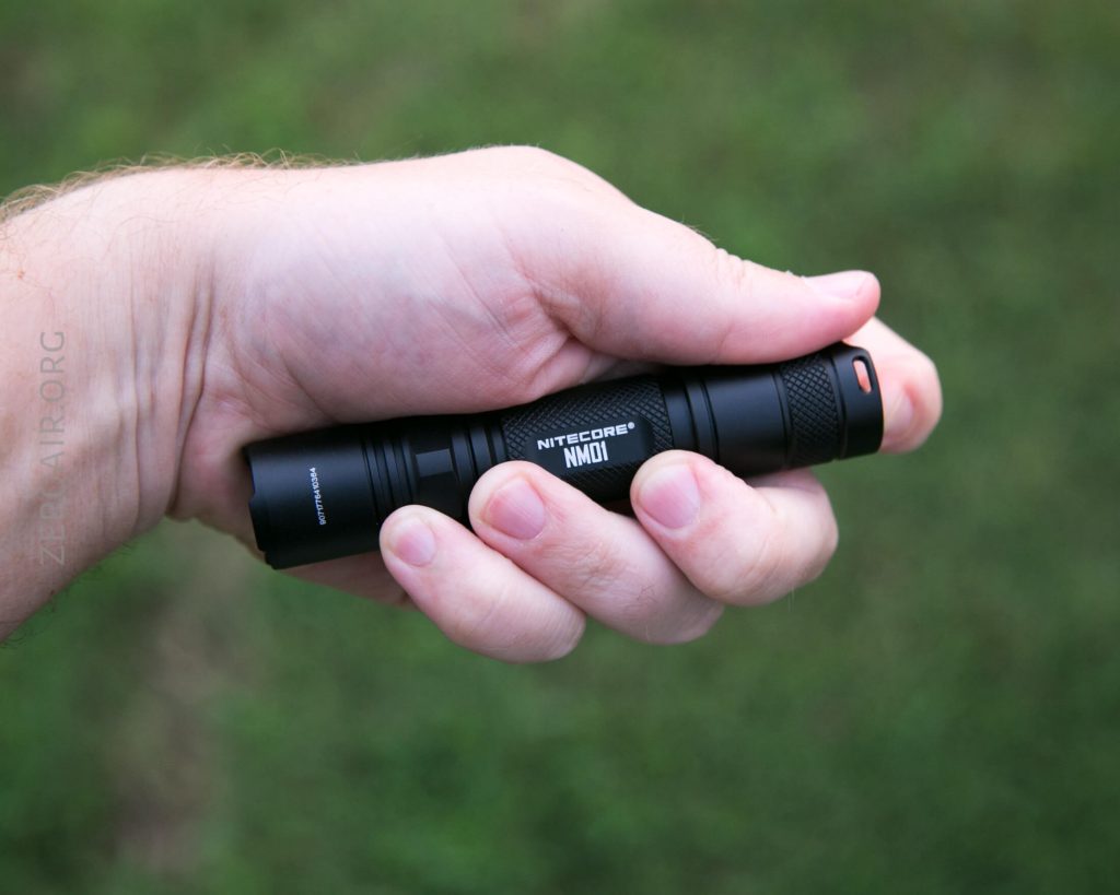 A persons hand holding a black Nitecore NM01 flashlight against a blurred green background.