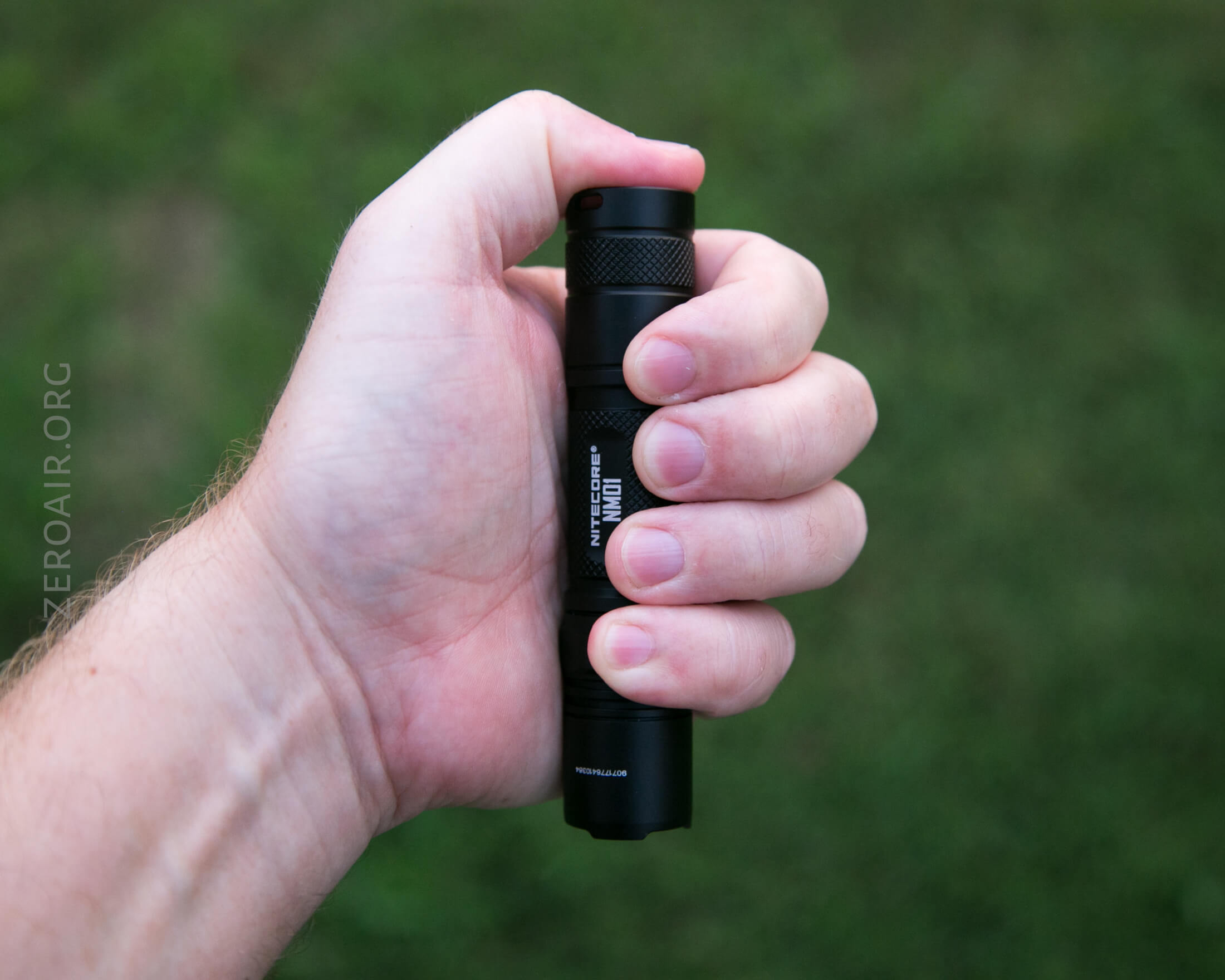 A hand holding a black flashlight outdoors with a grassy background, viewed from above. The flashlight is gripped firmly and text is visible on its side.