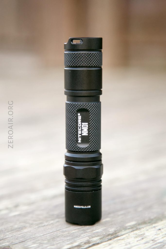 A black Nitecore NM01 flashlight stands upright on a wooden surface, with the brand name and model printed clearly on its side. The background is softly blurred.