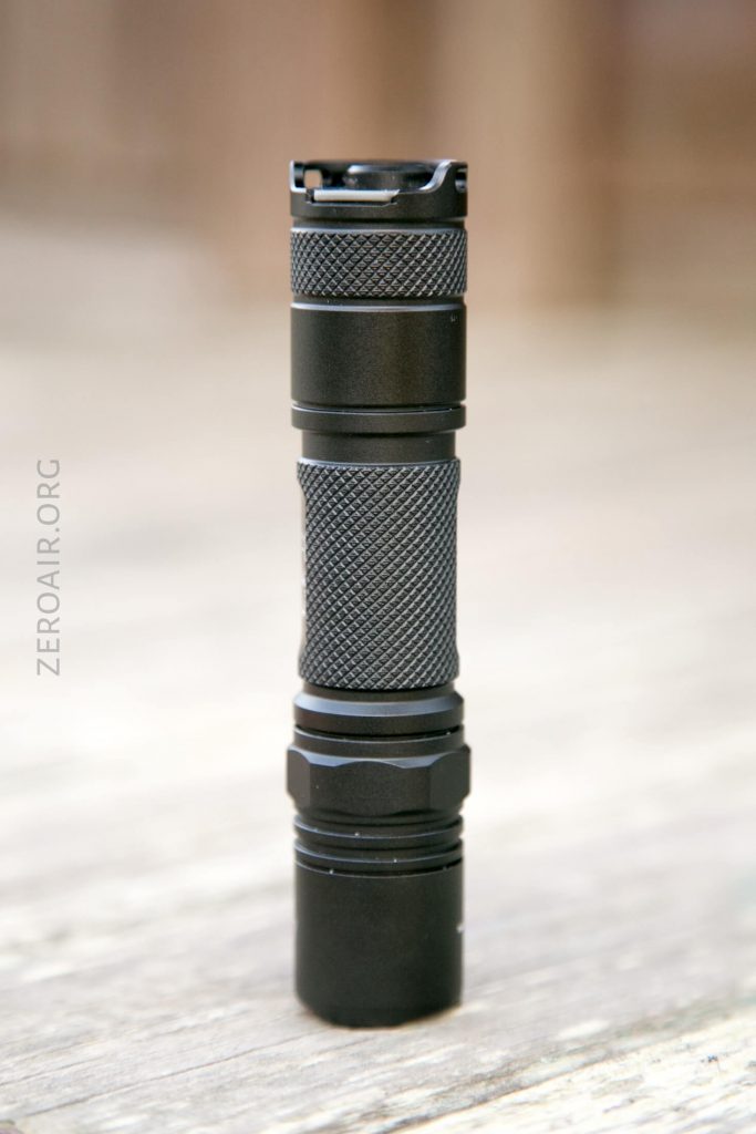 A compact black flashlight with a textured grip stands upright on a wooden surface. The background is blurred and the word ZEROAIR.ORG appears vertically on the left side.