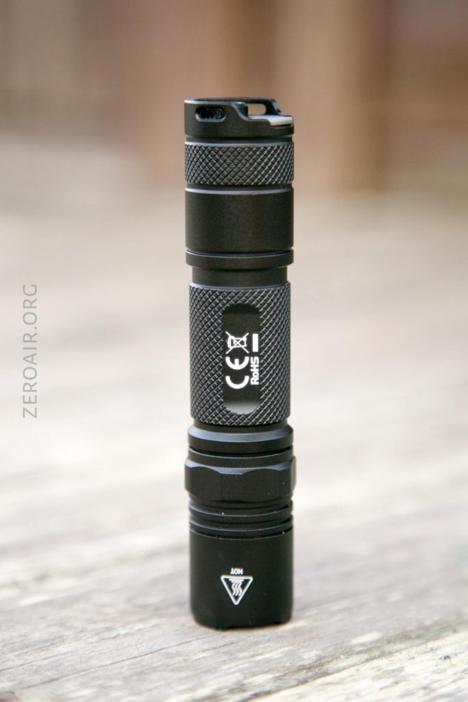 A small black flashlight stands upright on a wooden surface. The body has a textured grip, certification markings, and a symbol warning “hot” near the lens. The background is out of focus.